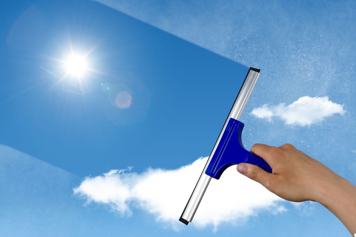 A person stands outdoors, holding a window cleaner high towards a clear blue sky, ready to clean windows. 