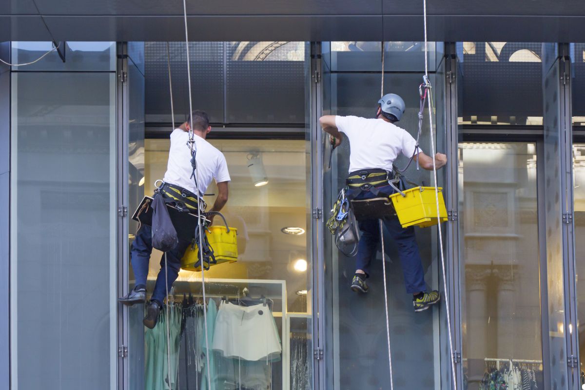 Two men using squeegees to clean windows on a tall building, with safety harnesses secured for protection.